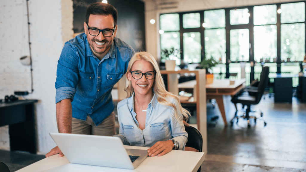 Dois profissionais sorrindo em um ambiente moderno de escritório. Um homem está em pé ao lado de uma mulher sentada, ambos usando roupas casuais, com um laptop aberto sobre a mesa. O ambiente possui grandes janelas com luz natural e móveis contemporâneos.