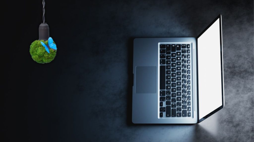 Notebook iluminado em fundo escuro simbolizando tecnologia, com lâmpada decorativa de musgo e borboleta azul representando sustentabilidade e impacto ambiental da inteligência artificial.