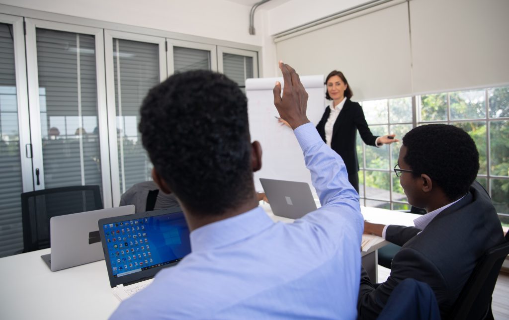 Profissionais em reunião de negócios em uma sala de conferências moderna; uma mulher de terno apresenta informações em um flip chart enquanto um dos participantes, sentado à mesa com um laptop aberto, levanta a mão para fazer uma pergunta.