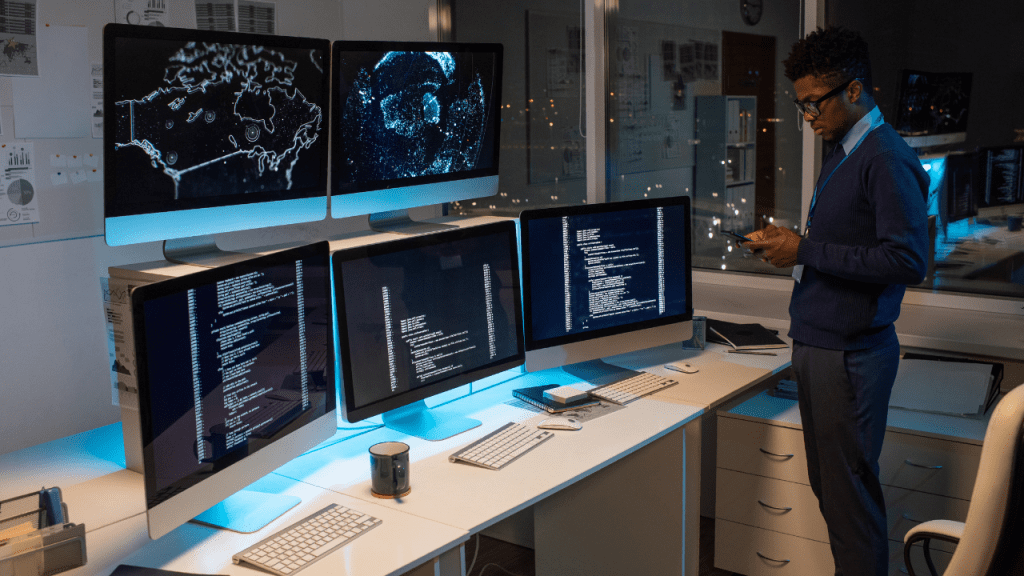 Profissional de TI em escritório noturno, diante de cinco monitores — três exibindo código e dois com mapas/visualizações — enquanto consulta o smartphone; cenário de monitoramento e cibersegurança.
