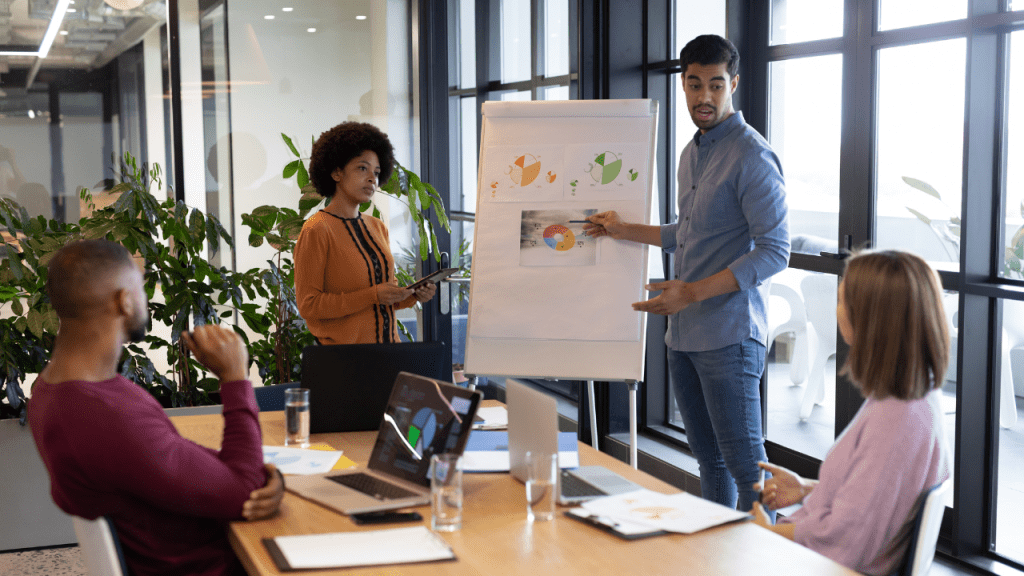 Equipe em reunião; apresentador aponta gráficos em um flip chart enquanto colegas acompanham com notebooks sobre a mesa em sala envidraçada.