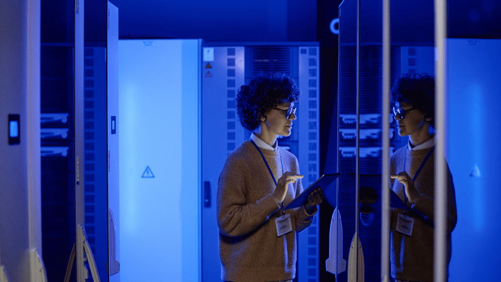 Profissional de TI com tablet inspeciona racks de servidores em data center azul, representando infraestrutura de IA e soberania digital na América Latina.