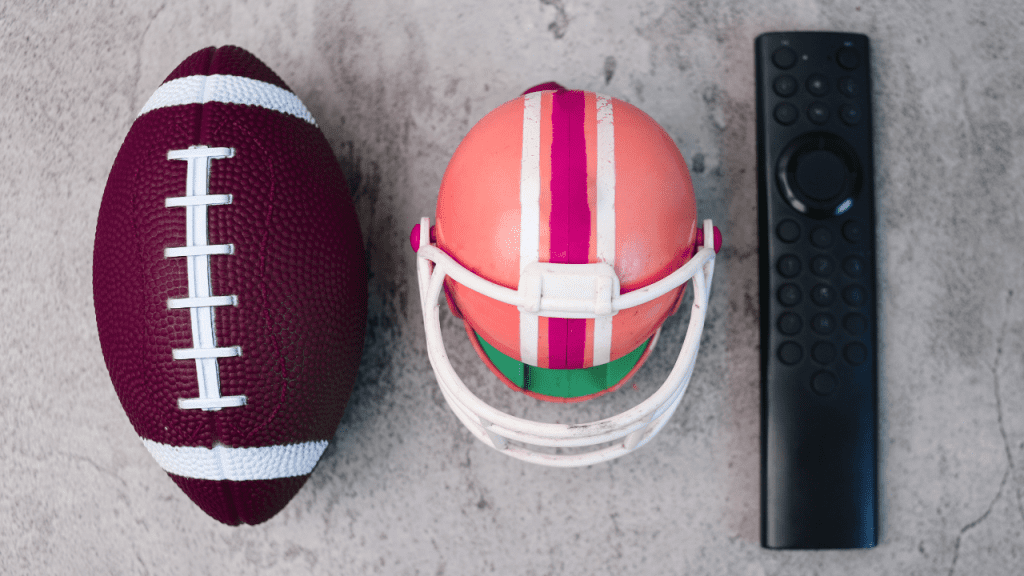 Bola de futebol americado ao lado de um capacete de jogo da cor rosa e vermelho e um controle de televisão, todos em fileira para trazer a ideias de conectividade futebol e tecnologia.