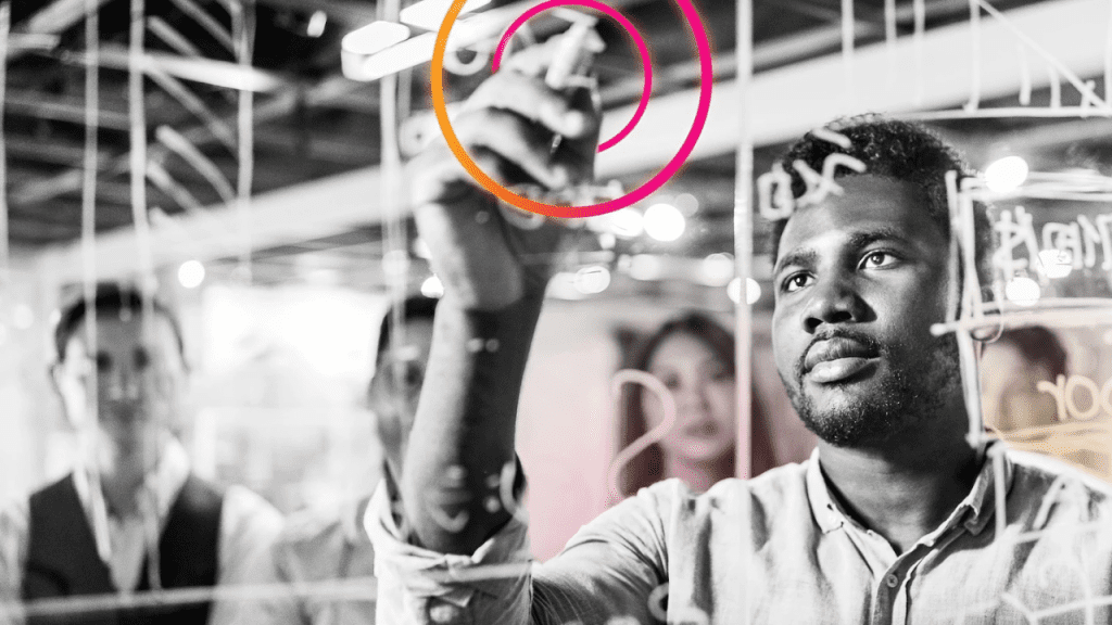 Profissional escrevendo com marcador em um quadro de vidro com diagramas; mão em destaque dentro de um círculo colorido, enquanto a equipe observa desfocada ao fundo em ambiente de escritório.