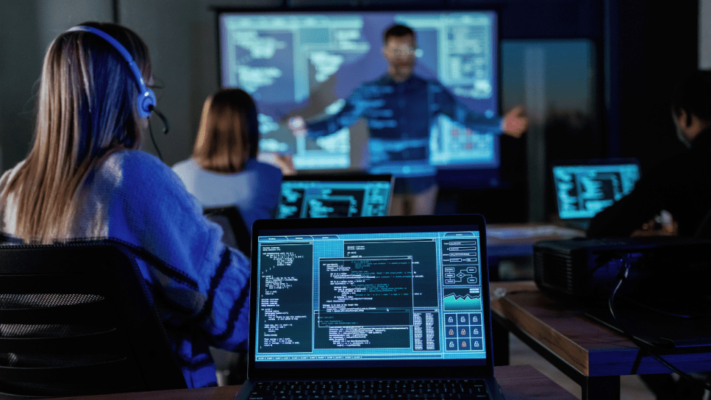 Treinamento de cibersegurança: notebook em primeiro plano exibe código e painéis enquanto um instrutor apresenta na tela projetada; participantes com headsets acompanham em sala de aula.