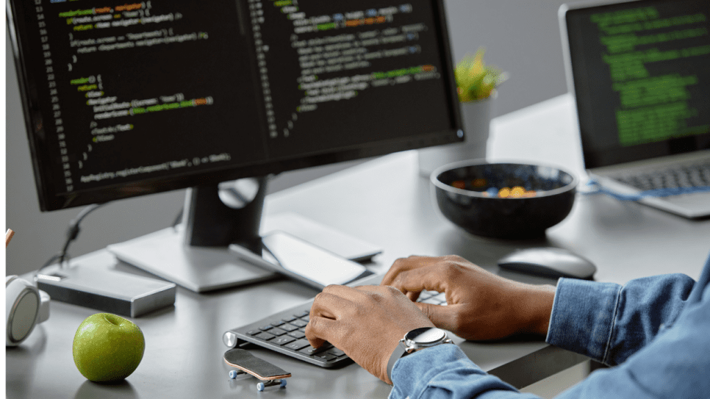 Pessoa desenvolvedora de software digitando em um teclado em frente a dois monitores com código na tela, em um ambiente de escritório, representando o dia a dia da programação.