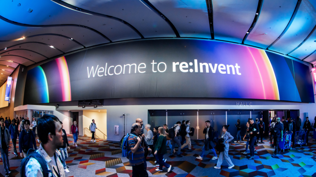 Corredor de entrada do evento AWS re:Invent com grande painel iluminado exibindo a frase “Welcome to re:Invent”, pessoas caminhando em direção aos salões e clima movimentado de conferência de tecnologia em Las Vegas.
