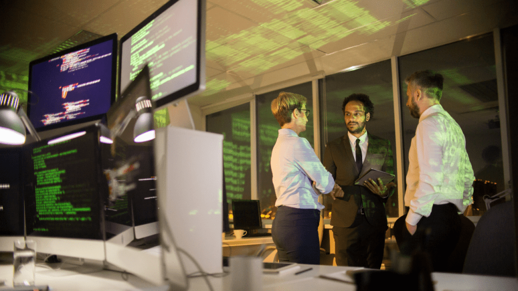 Três profissionais em um escritório à noite discutem estratégia de cibersegurança enquanto telas exibem linhas de código e reflexos verdes, sugerindo monitoramento e proteção de dados corporativos.