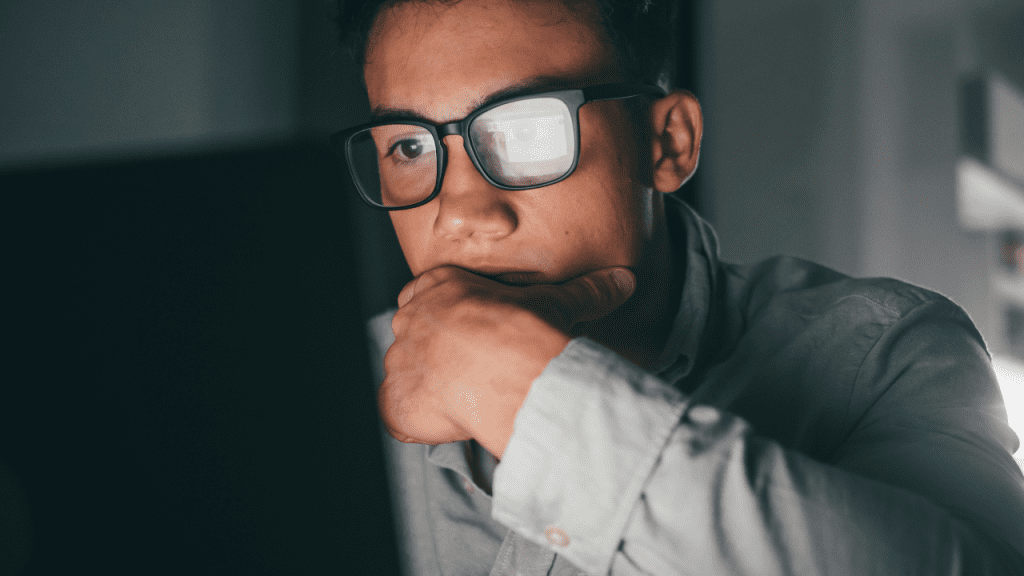 Profissional de TI com óculos, em ambiente com pouca luz, observa atentamente uma tela e apoia a mão no queixo, sugerindo análise de segurança e monitoramento de rede.
