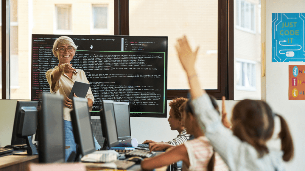Professora em sala de aula de programação aponta para alunos com a mão levantada, enquanto um painel ao fundo exibe linhas de código, representando educação em tecnologia e aprendizado com IA.