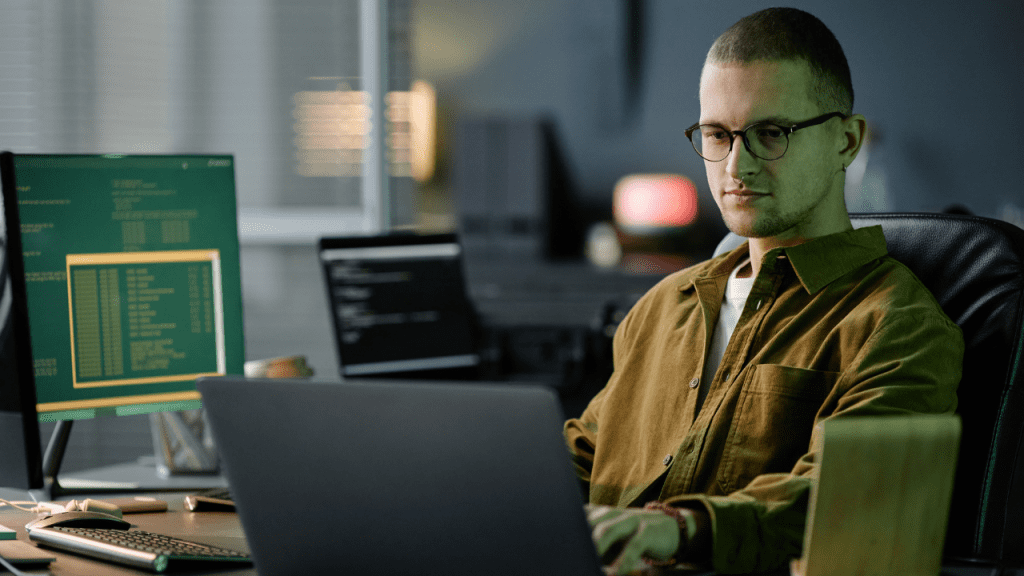 Homem usando óculos trabalha em um notebook em ambiente de escritório com iluminação baixa, enquanto monitores ao fundo exibem linhas de código e interfaces de programação.