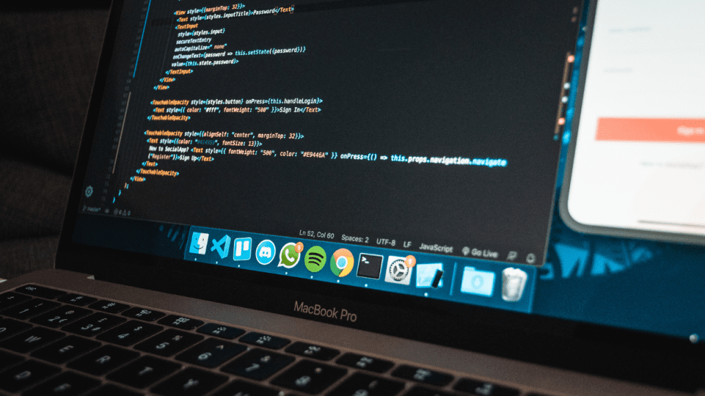 Close de um MacBook Pro exibindo código em JavaScript na tela, com interface de desenvolvimento aberta e elementos de aplicativo ao fundo, representando programação, upskilling e reskilling em TI.