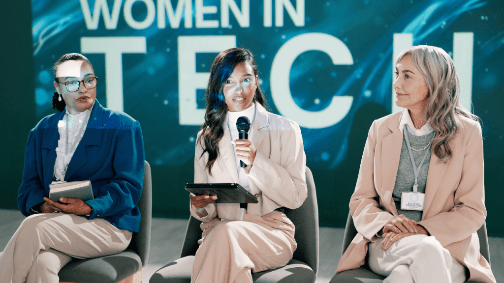 Mulheres participando de um painel sobre tecnologia, com três profissionais sentadas em cadeiras no palco; a mulher ao centro segura um microfone e um tablet enquanto fala, com o fundo exibindo o texto “Women in Tech”, representando liderança feminina e presença de mulheres na área de tecnologia.