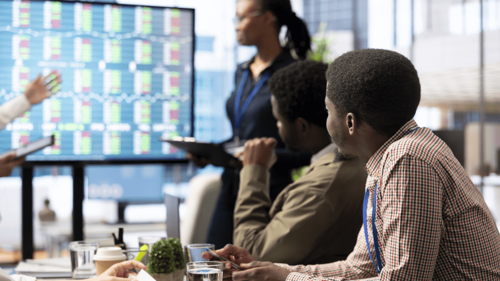 Equipe em reunião corporativa observa um grande painel com gráficos e dados de mercado, enquanto uma pessoa apresenta informações na tela e os participantes acompanham com tablets e anotações em um ambiente de escritório.