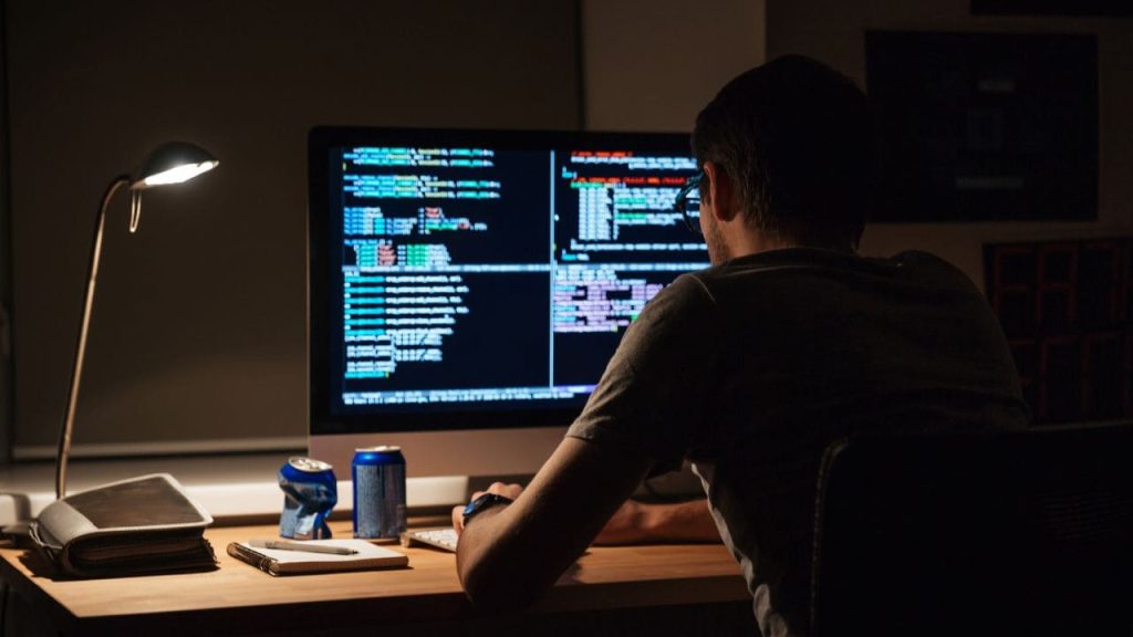 Profissional analisando códigos em um computador durante a noite, representando ataques hackers com IA, cibersegurança corporativa e proteção de dados empresariais.