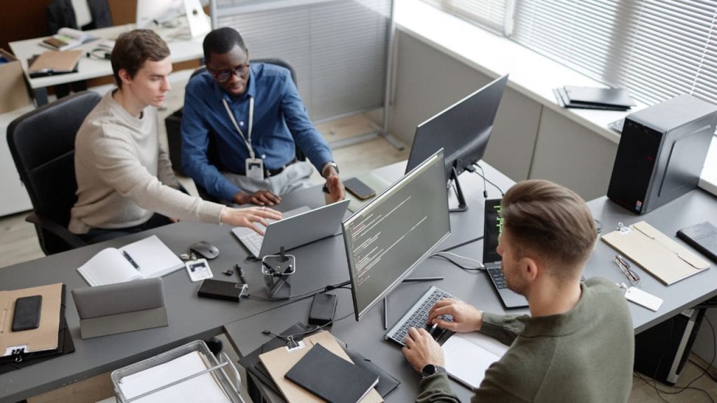 Profissionais de TI trabalhando em equipe em uma sala corporativa, com computadores e monitores exibindo códigos, representando cibersegurança, tecnologia e capacitação profissional.