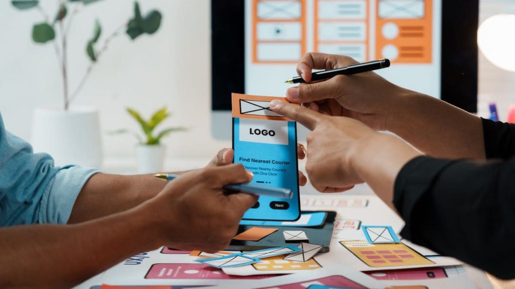 Profissionais desenham protótipo de aplicativo em smartphone, representando o uso de tecnologia e inteligência artificial para melhorar a segurança da experiência digital.