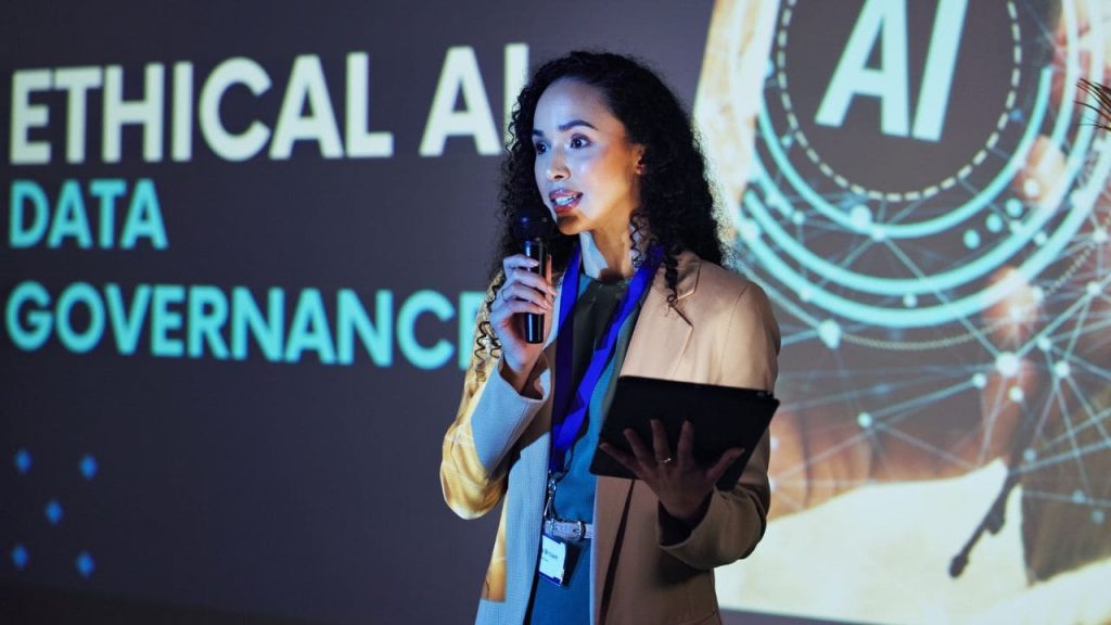 Mulher palestrando em um evento sobre inteligência artificial, dados e governança, segurando um microfone e um tablet diante de um telão com os dizeres “ETHICAL AI”, “DATA” e “GOVERNANCE”.