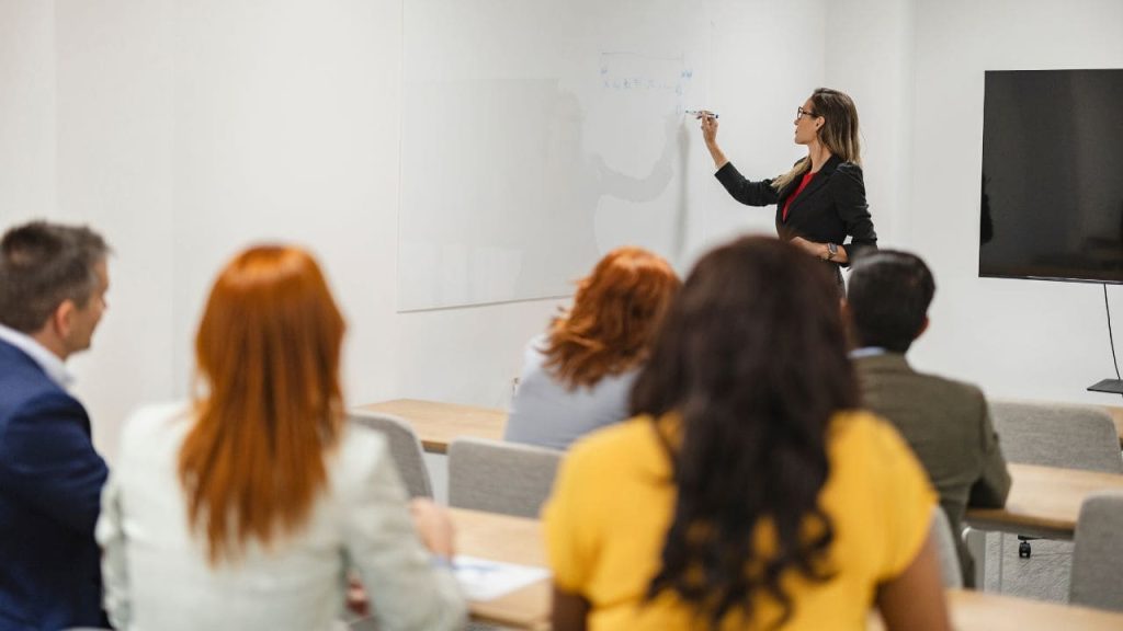 Mulher ministrando uma aula ou treinamento corporativo, escrevendo em um quadro branco enquanto profissionais acompanham a apresentação em uma sala de treinamento.