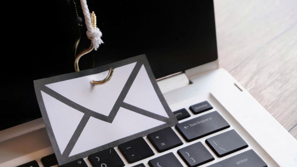 Envelope de papel fisgado por um anzol sobre o teclado de um notebook, representando um ataque de phishing por e-mail para roubo de credenciais.