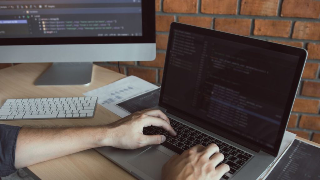 Desenvolvedor digitando código em um notebook, com um segundo monitor exibindo código ao fundo, representando a revisão de consultas ao banco de dados para prevenir injeção de SQL com prepared statements.
